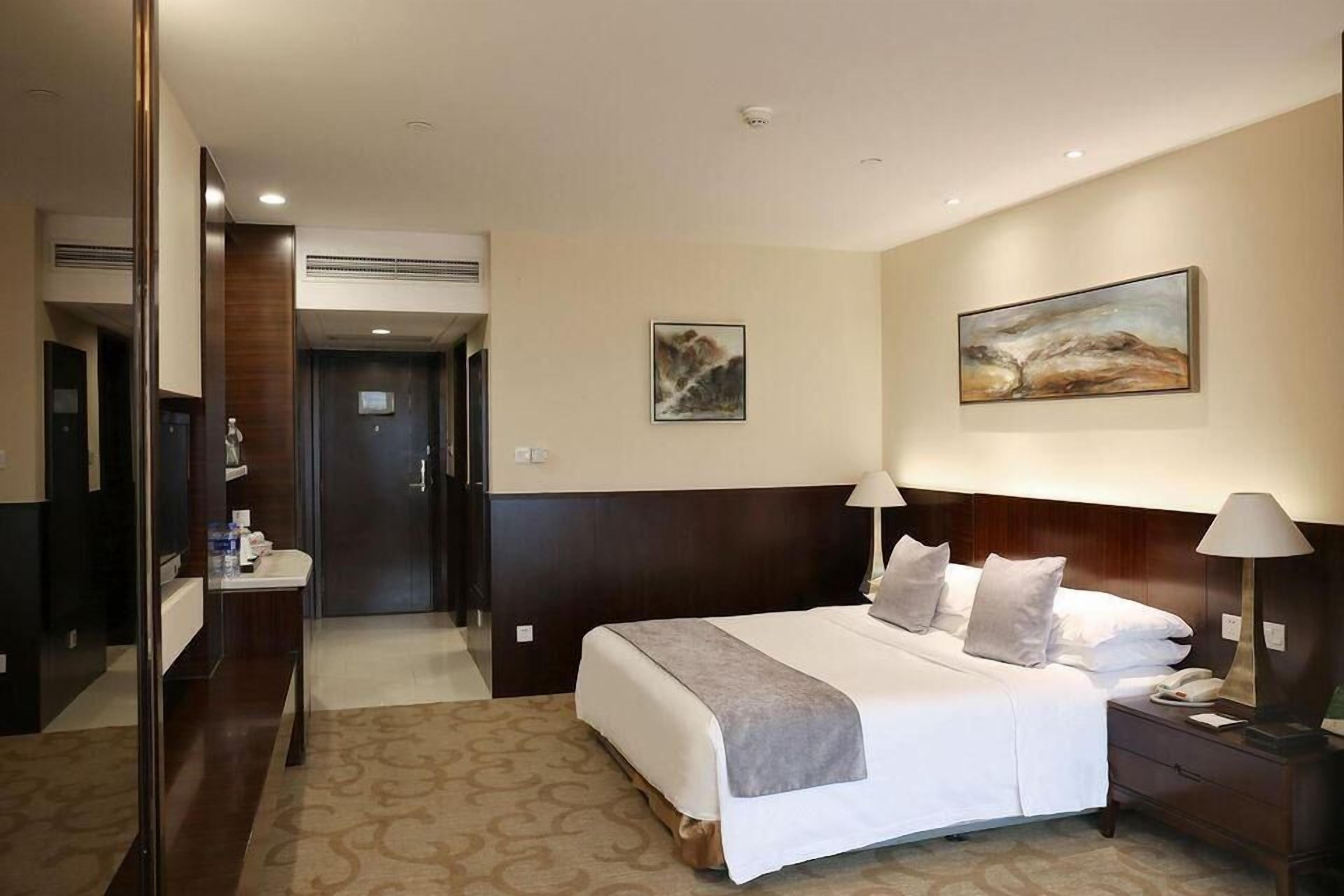 Een hotelkamer met een groot bed, bedlampjes, een nachtkastje, een spiegel, een schilderij aan de muur en een deur die naar een andere kamer leidt.