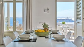Uma mesa de jantar com uma tigela de fruta, cadeiras brancas e vista para o mar através de grandes janelas.