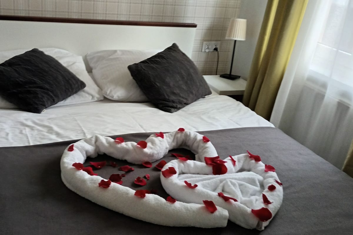Una habitación de hotel con una cama bien hecha, dos mesitas de noche con lámparas y vistas al exterior a través de una ventana con cortinas.