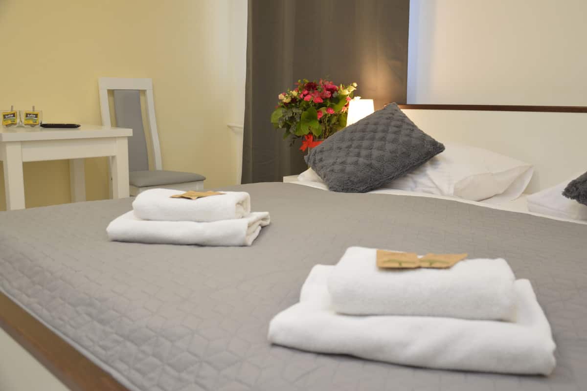 Una cama bien hecha con toallas blancas y una almohada gris, una mesita con un control remoto y un jarrón con flores.