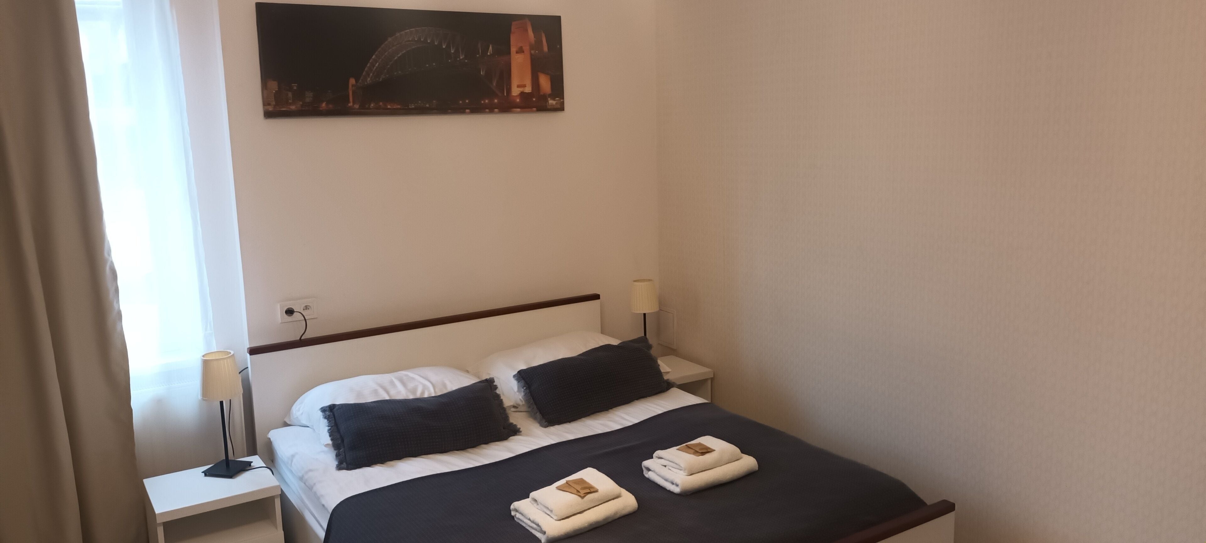 Une chambre d’hôtel avec un lit, deux oreillers, une table de chevet avec une lampe et un tableau représentant un pont accroché au mur.