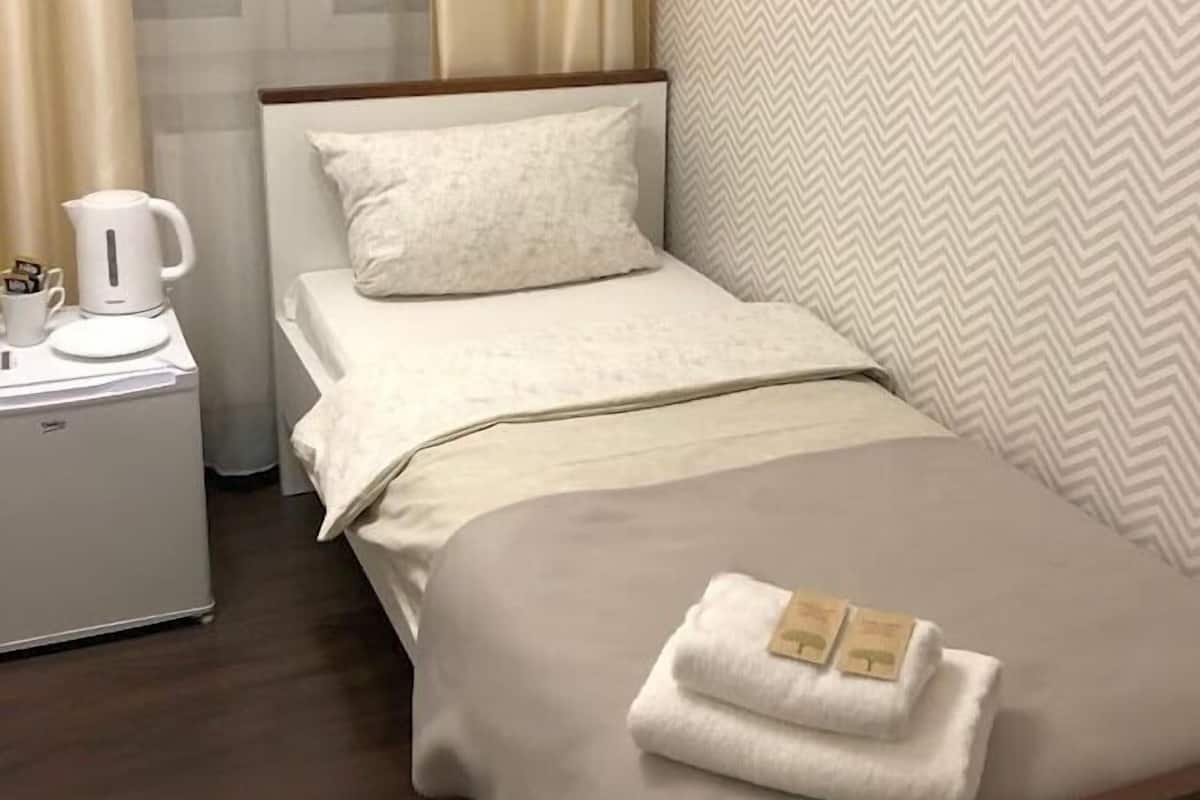 Una cama pequeña y bien hecha con edredón blanco y manta marrón, una mesita de noche con hervidor blanco y una taza, y una pared con diseño en zigzag.