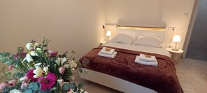 Una cama bien hecha con cubierta marrón, dos almohadas blancas y dos toallas dobladas a un lado. Se observa una mesita de noche con una lámpara y un pequeño objeto decorativo. Un arreglo floral grande es visible en primer plano.