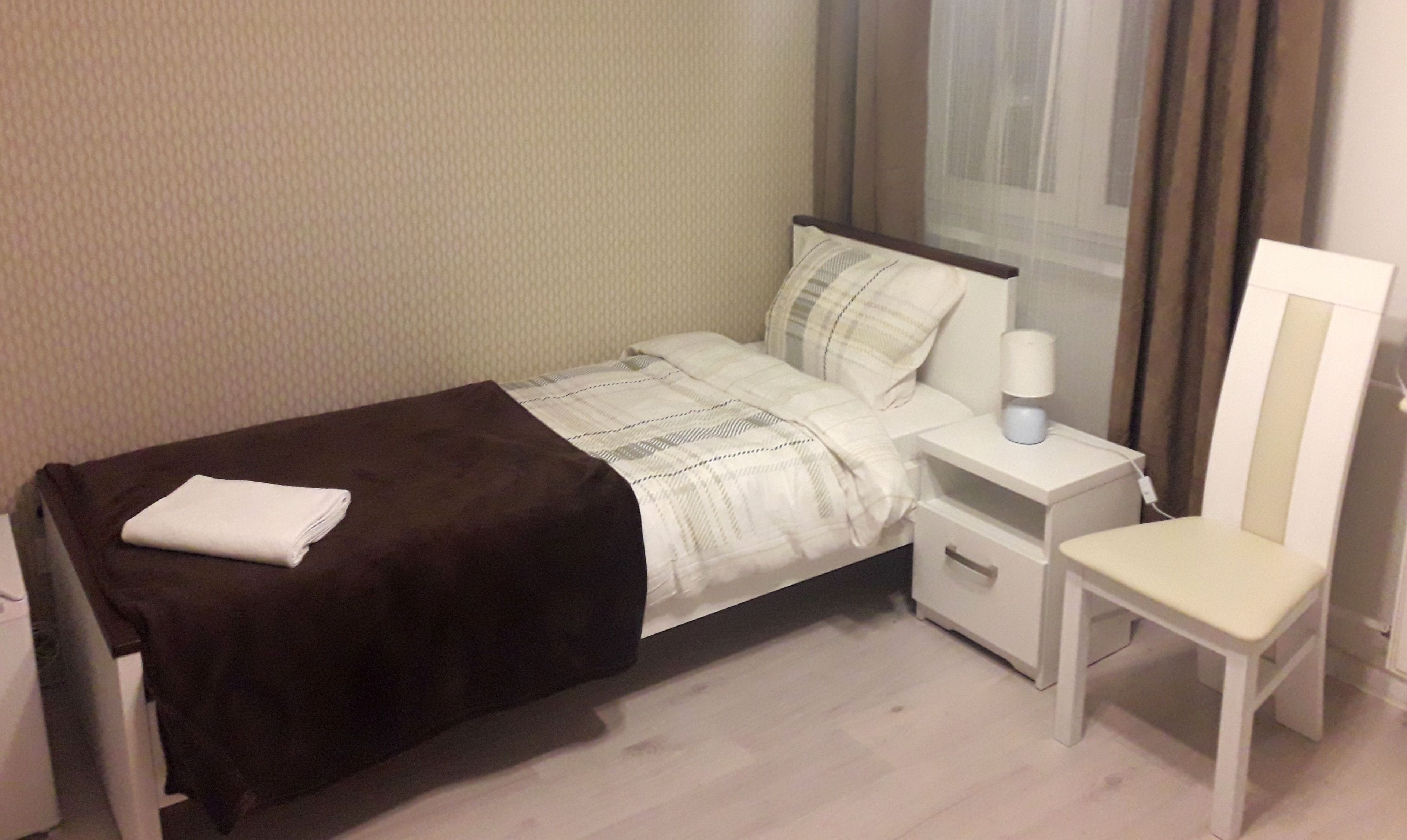 Cama bem arrumada com cobertura escura, mesa de cabeceira com abajur e uma cadeira.