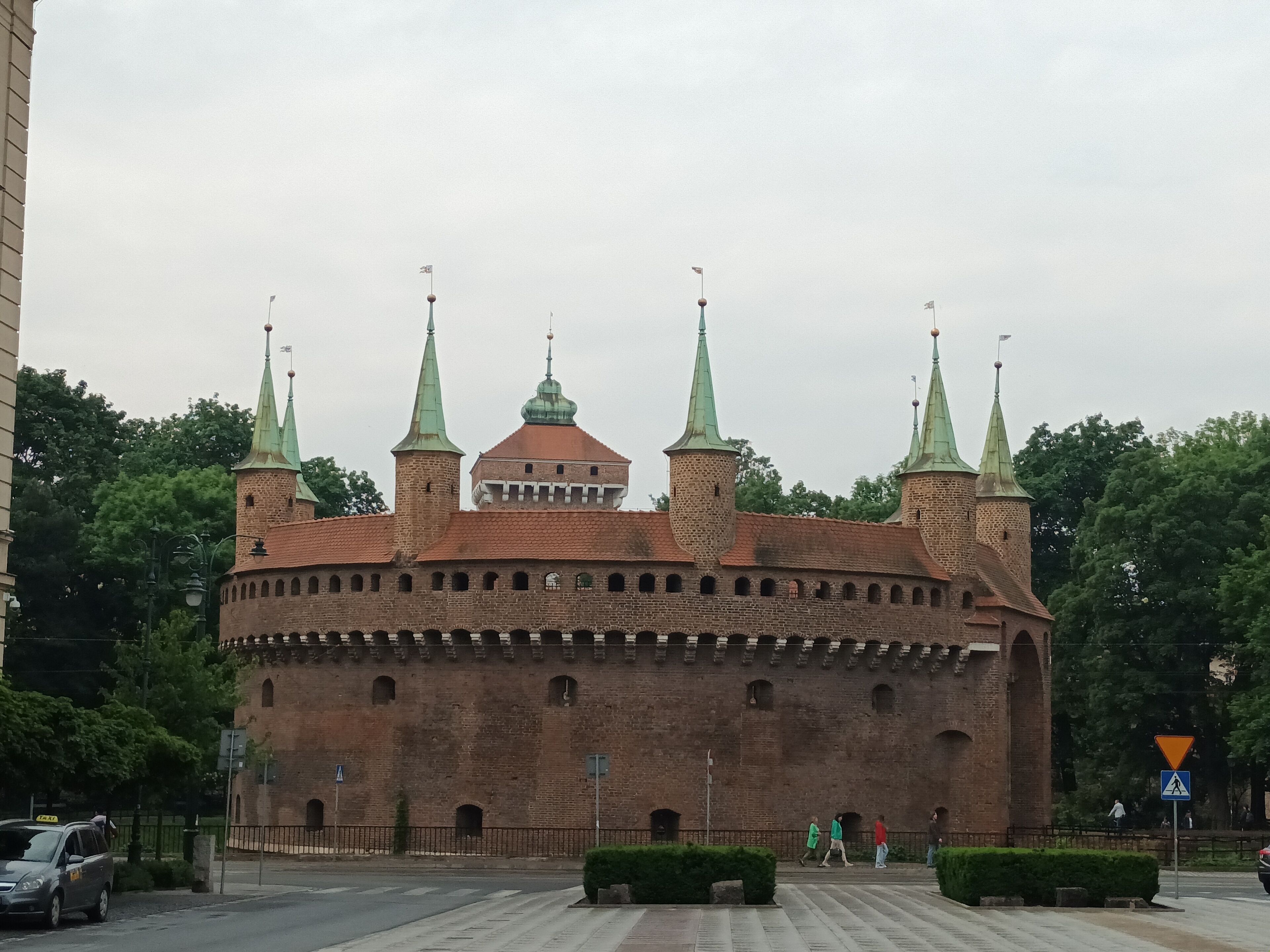 A historic brick building with multiple towers and a central dome, surrounded by greenery and a paved area.