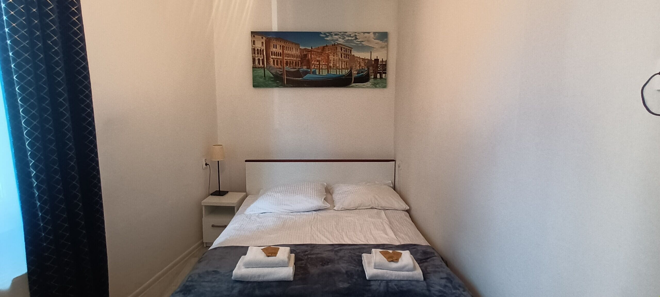 A narrow hotel room with a bed, a nightstand, a lamp, and a painting of a canal scene.