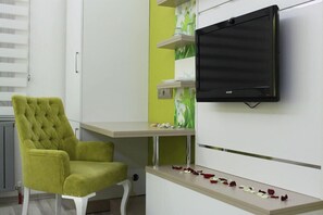A room with a green tufted chair, a flat-screen TV mounted on the wall, and a shelf with decorative items. - Derya Otel (Konya)