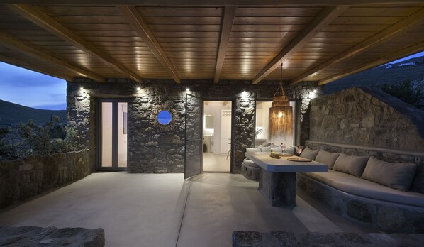 Un patio al aire libre con paredes de piedra, techo de madera, un banco de piedra y una mesa con vasos y un bol.