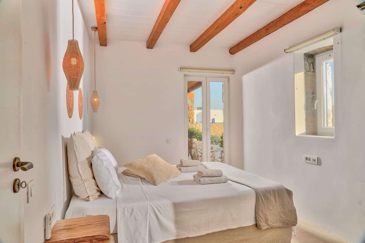 A bedroom with a bed, pillows, a bedside table, a hanging light fixture, and a window with a view.