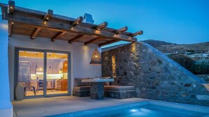 A modern outdoor pool area with a stone wall and wooden pergola. - VLIA Myconian Residences (Mykonos)