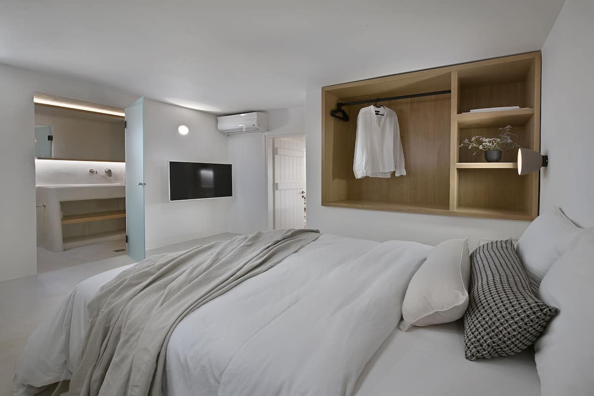A bedroom with a bed, a TV, a wooden shelf with clothes, and a bathroom visible through an open door.