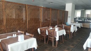 A restaurant with wooden lattice partitions, tables with white tablecloths, and a kitchen area in the background.