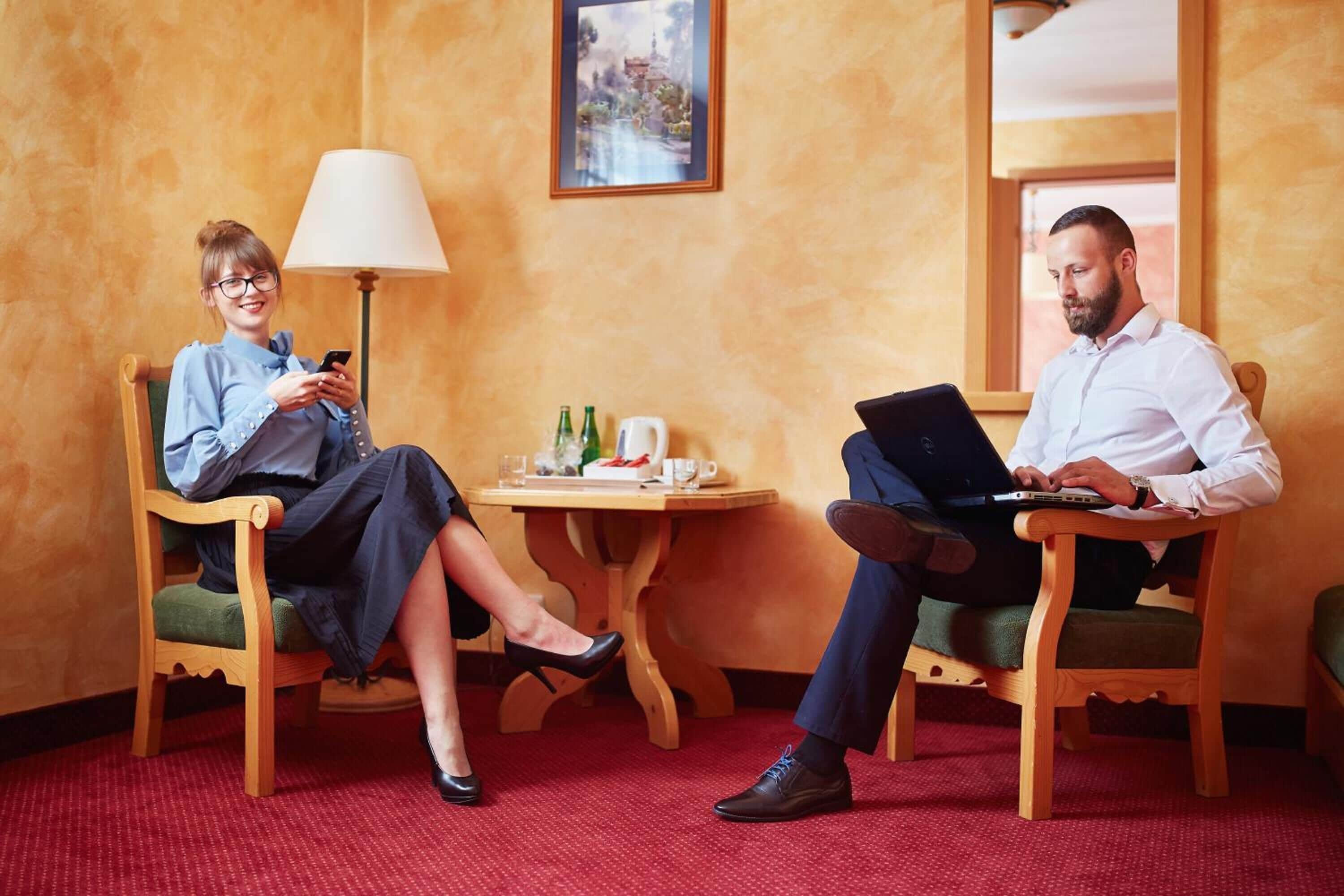 Twee personen zitten in een kamer met houten stoelen, een klein tafeltje met drankjes en een lamp.