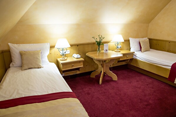 Habitación de hotel con dos camas individuales, una mesita de noche de madera con lámpara, una pequeña mesa con un jarrón y una alfombra roja.