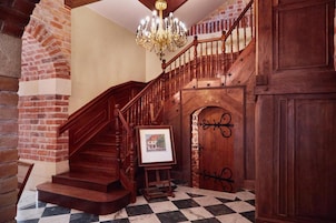 Uma escadaria imponente com corrimão de madeira e um candelabro, levando a uma porta com portão de ferro decorativo.
