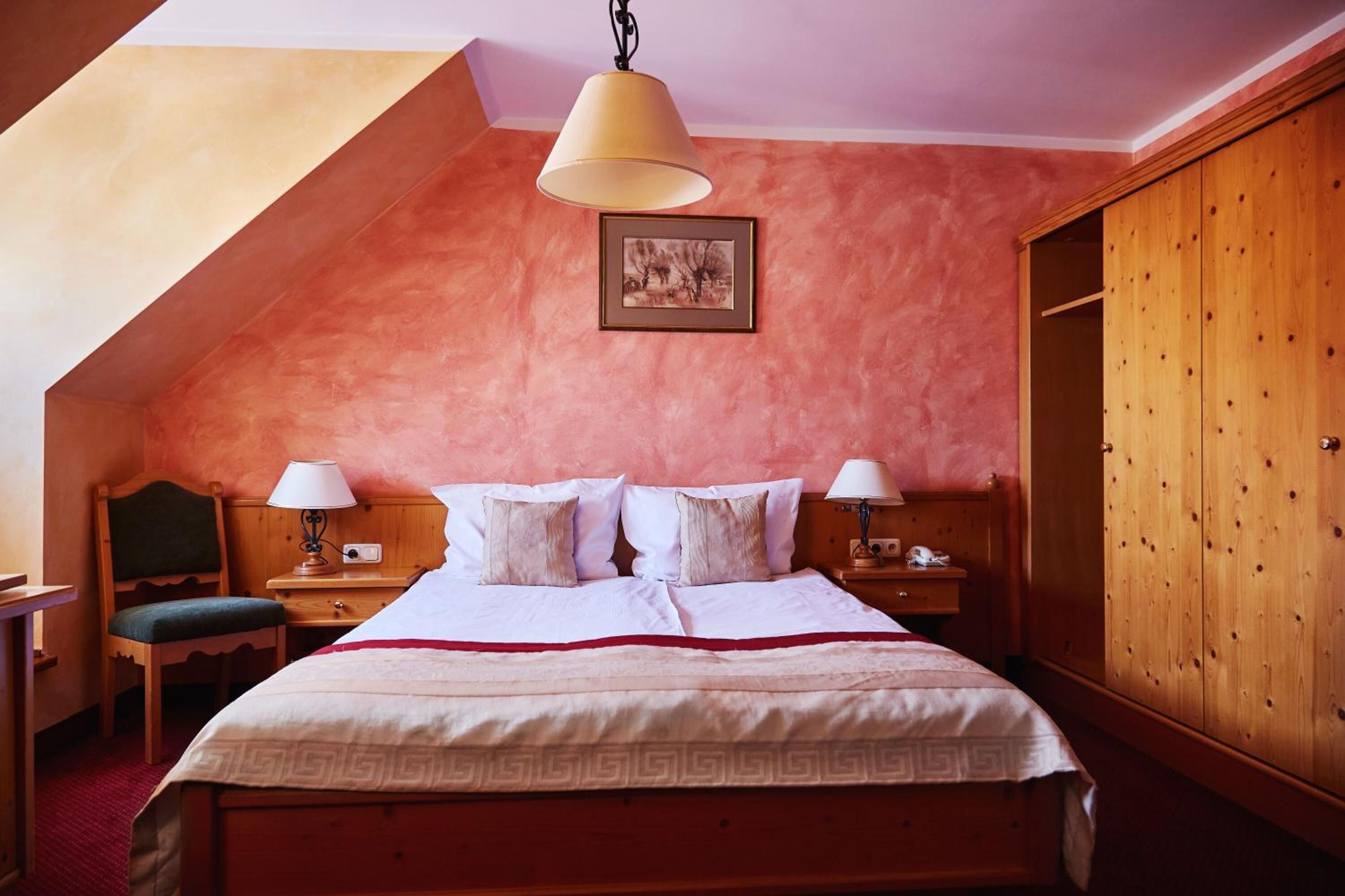 Un dormitorio con techo inclinado, una cama con sábanas blancas, dos lámparas de noche, un armario de madera y un cuadro enmarcado en la pared.