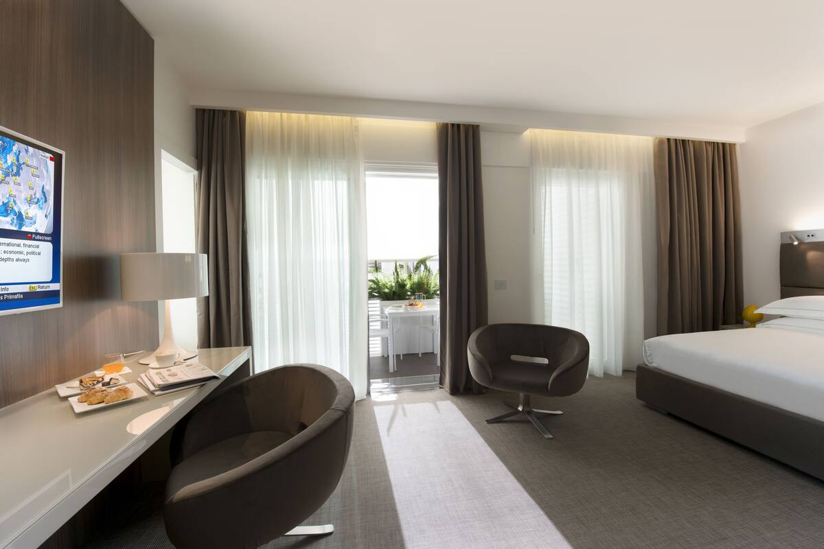 A hotel room with a bed, a desk with a chair, a small table with a chair, and a view of the outdoors through the window.