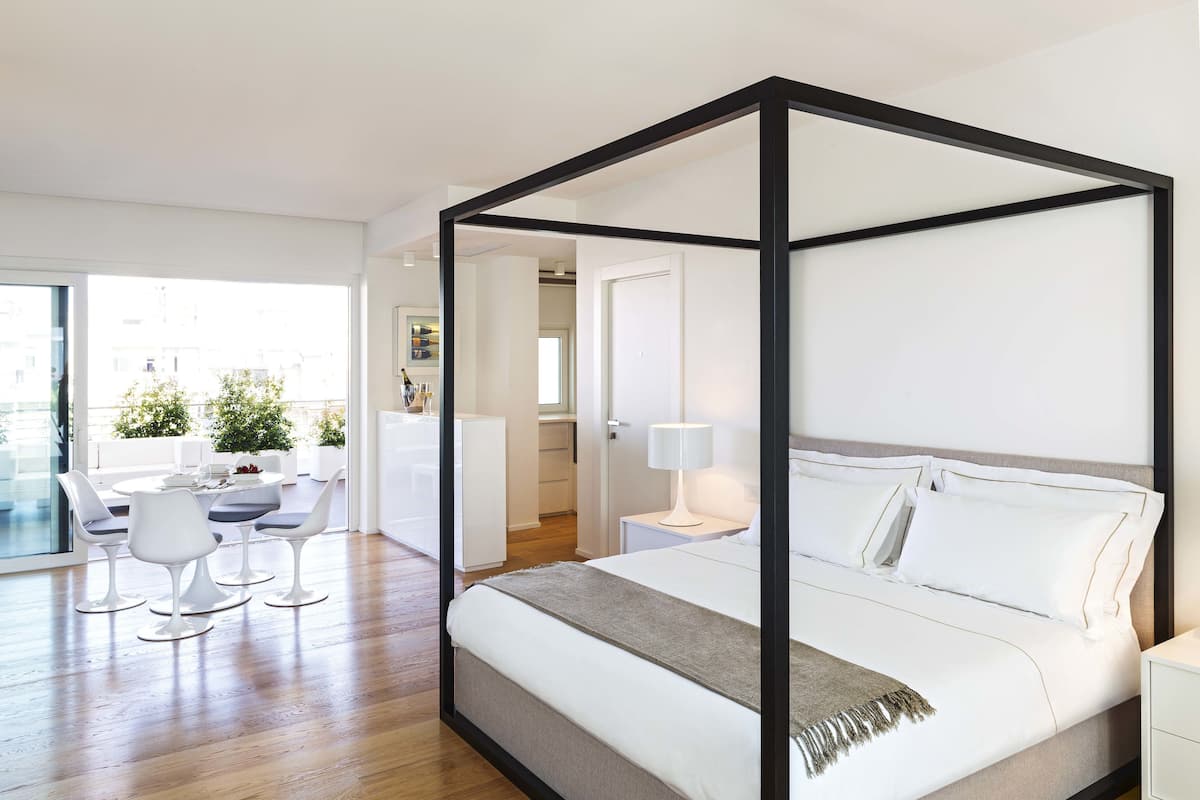 A modern bedroom with a four-poster bed, a dining area with white chairs, and a view of the city through large windows.