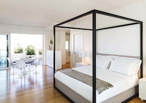 Un dormitorio moderno con cama con dosel, una zona de comedor con sillas blancas y vistas a la ciudad a través de amplios ventanales.
