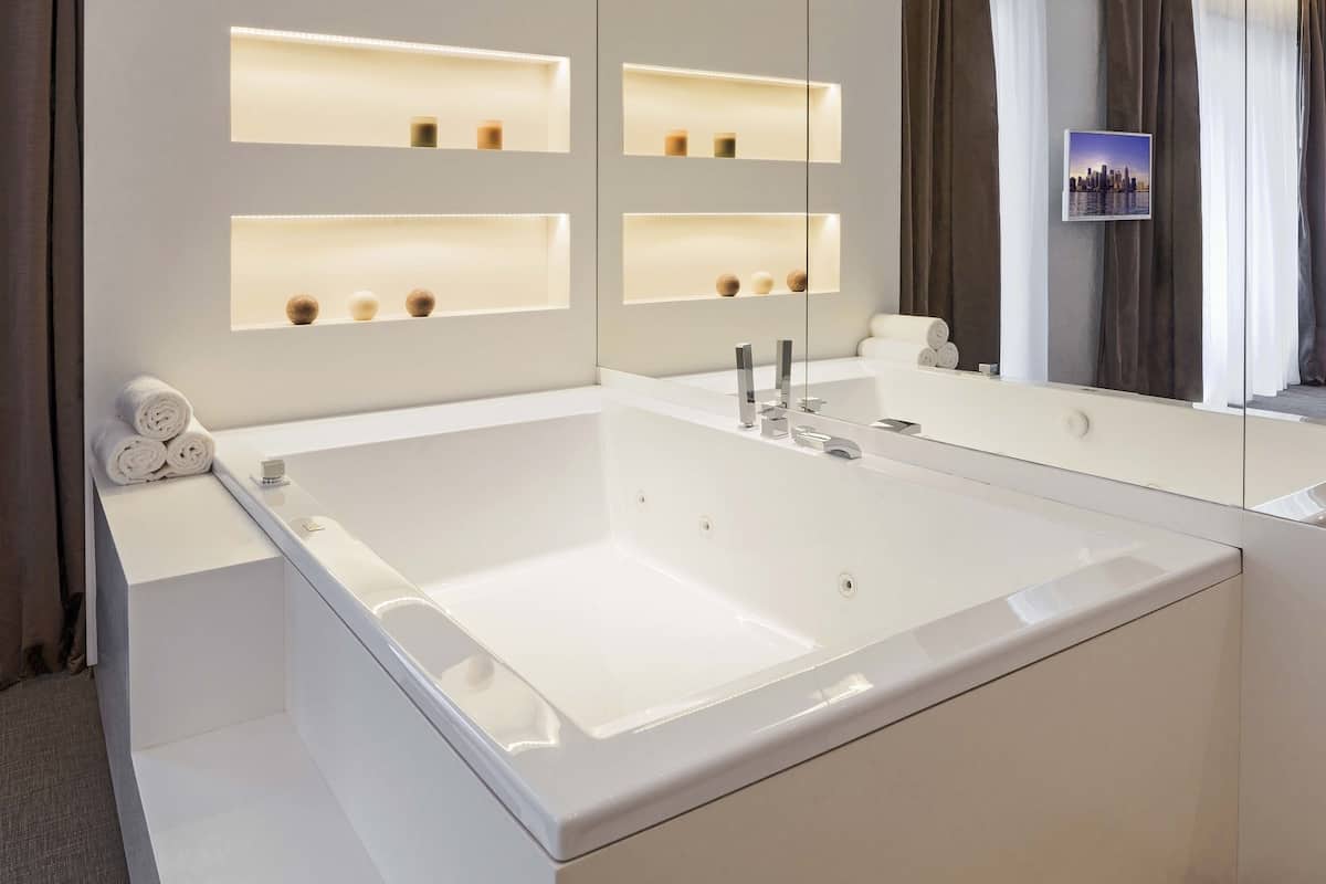 A modern bathroom with a large bathtub, built-in shelves with candles, and a cityscape photo on the wall.