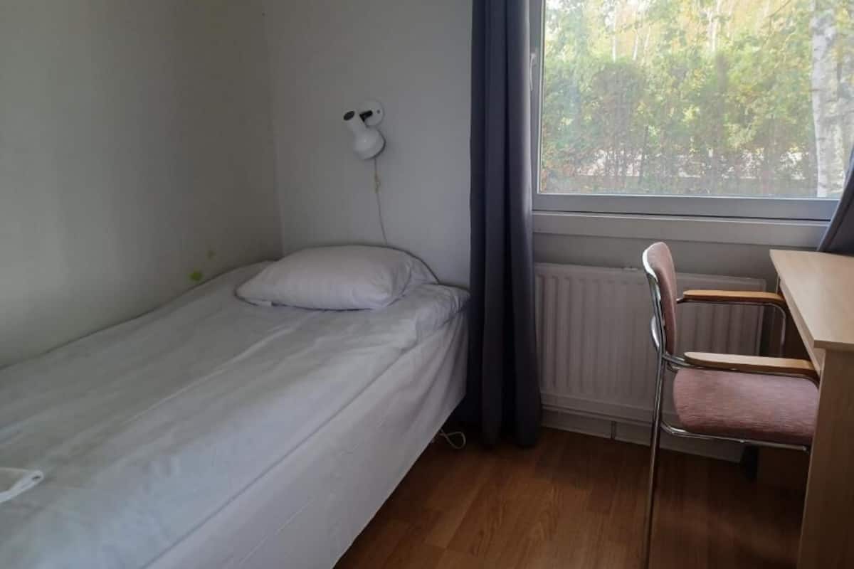Une petite chambre avec un lit simple, un plancher en bois, un bureau avec une chaise, un radiateur et une fenêtre avec des rideaux.