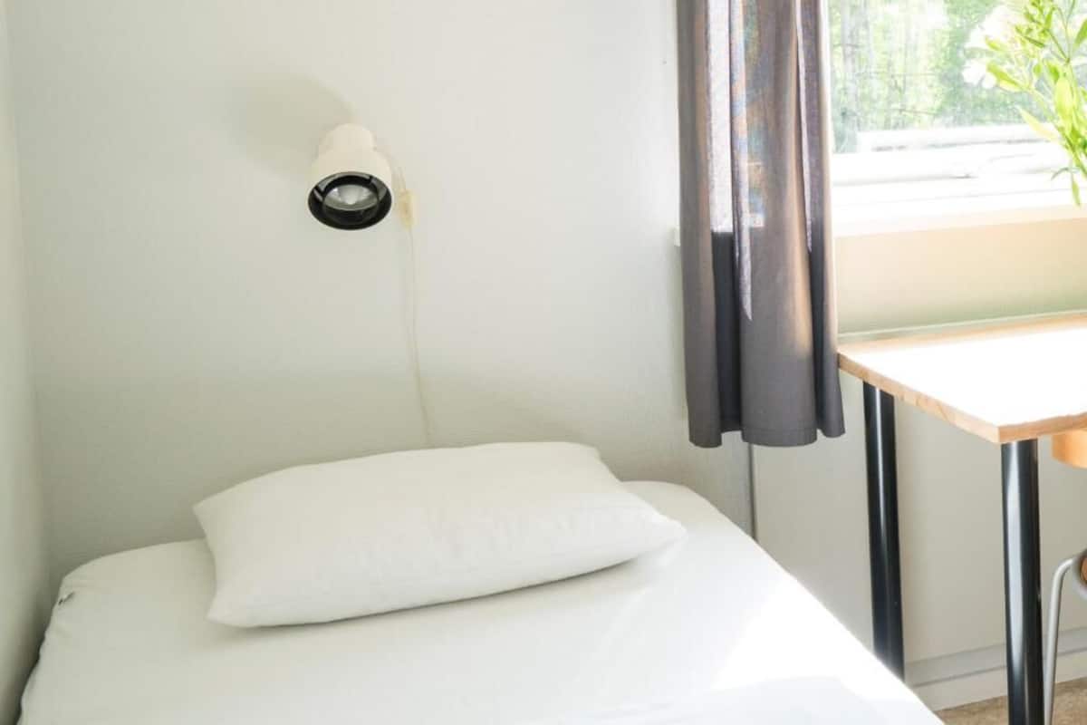 Un petit lit bien fait, avec un oreiller blanc et une lampe fixée au mur.