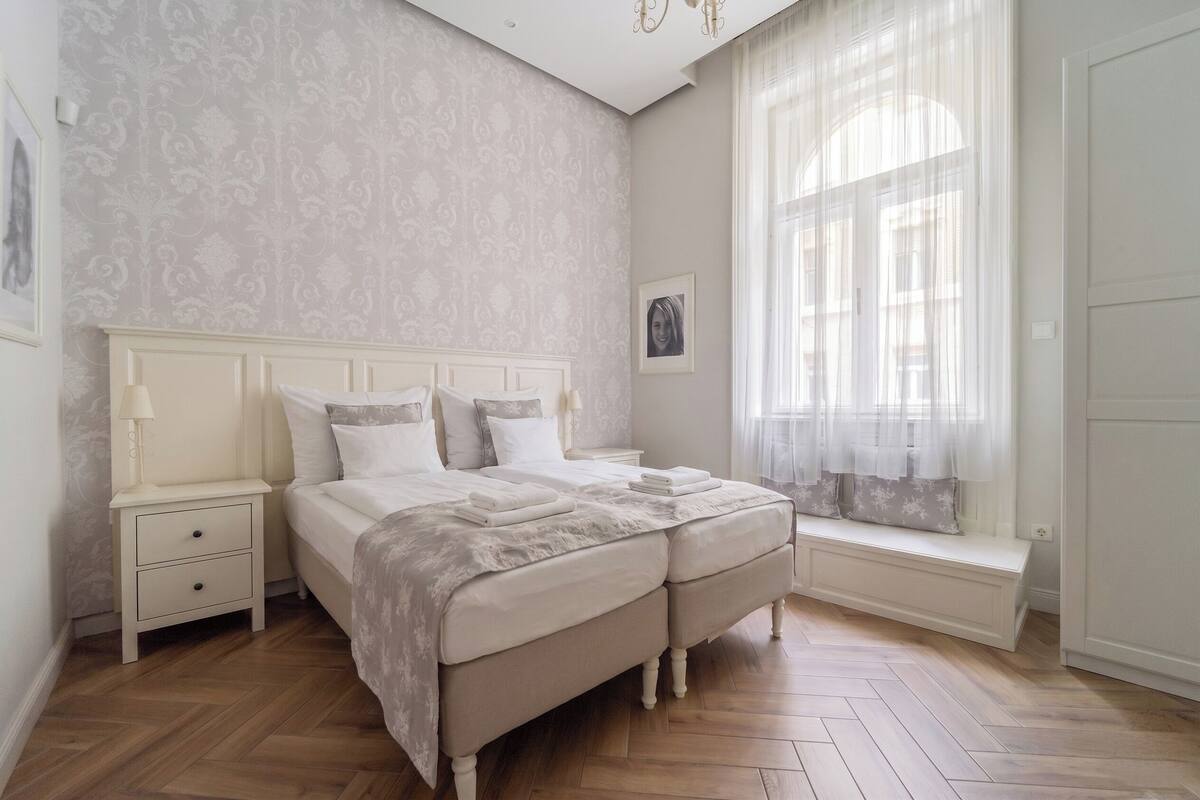 A bedroom with a bed, nightstand, bench, and window with curtains.