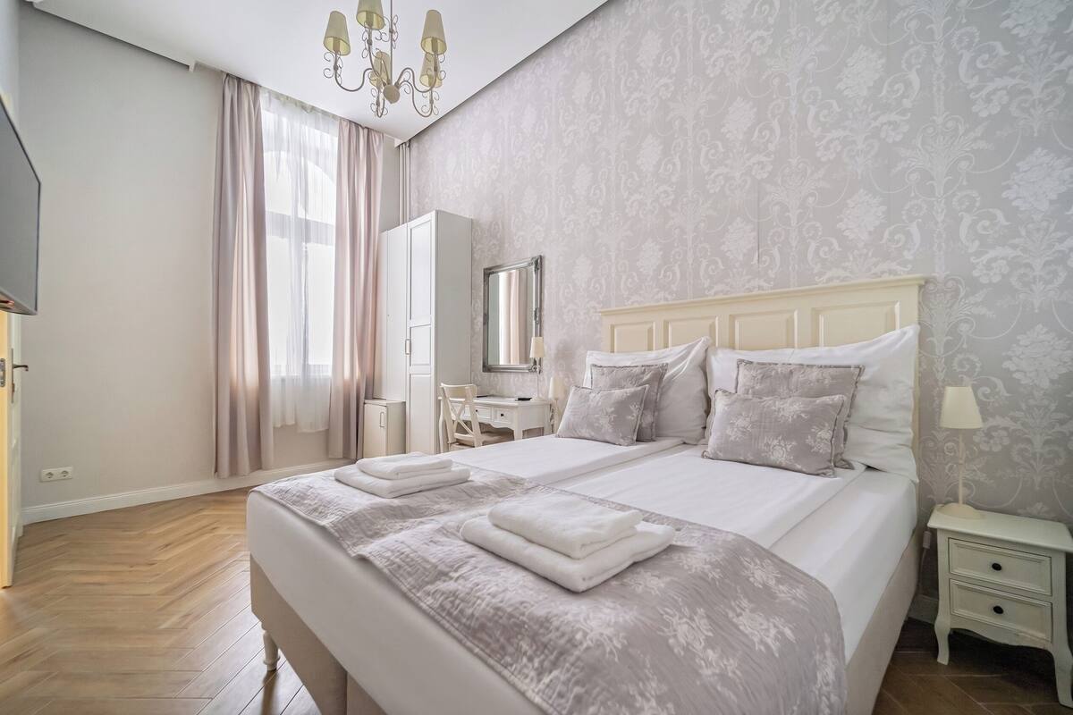 A double bed with a white headboard, a bedside table, a mirror, and a wardrobe in a room with patterned wallpaper and wooden flooring.