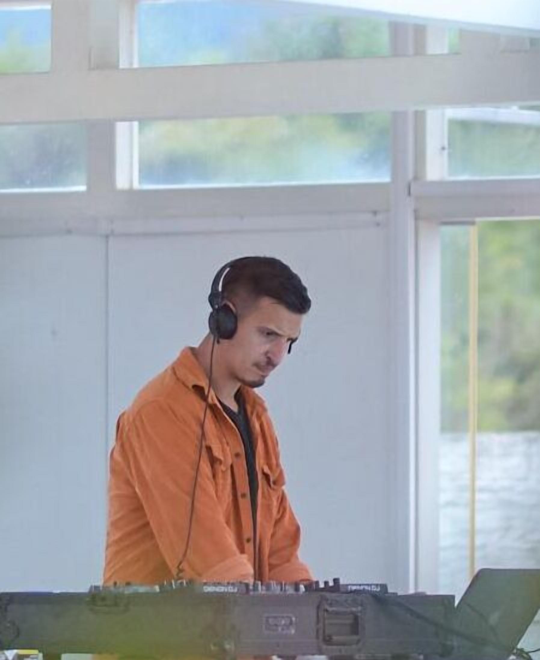 A person in an orange shirt standing in front of a DJ setup with a laptop and a mixer.