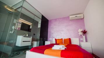 A modern hotel room with a glass shower enclosure, a bed with red and orange bedding, and a white nightstand with a vase of flowers.