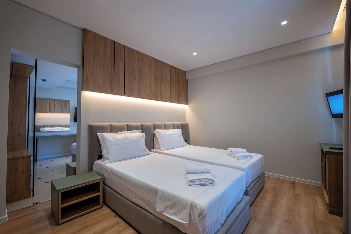 A modern bedroom with two beds, wooden paneling above the beds, a small bedside table, and a flat-screen TV on the wall.