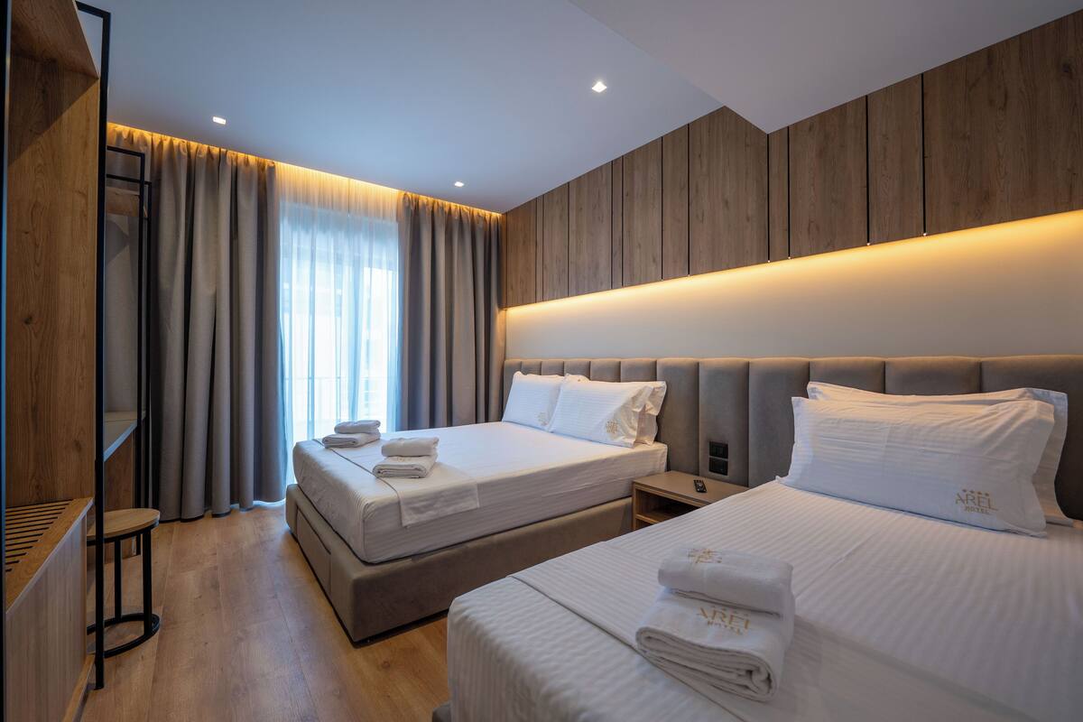 A modern hotel room with two beds, wooden panel walls, and a large window with curtains.