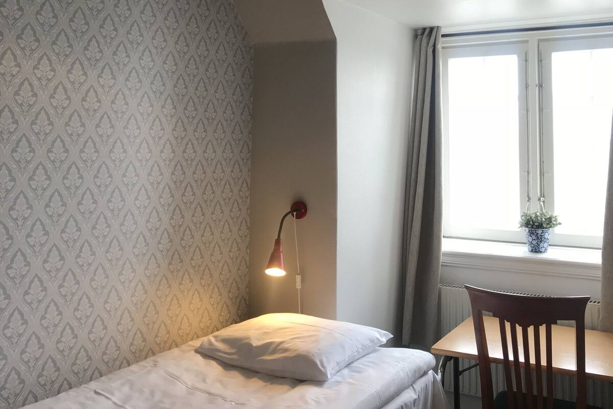 A small, neatly arranged room with a single bed, a table with a chair, a hanging lamp, and a window with curtains.