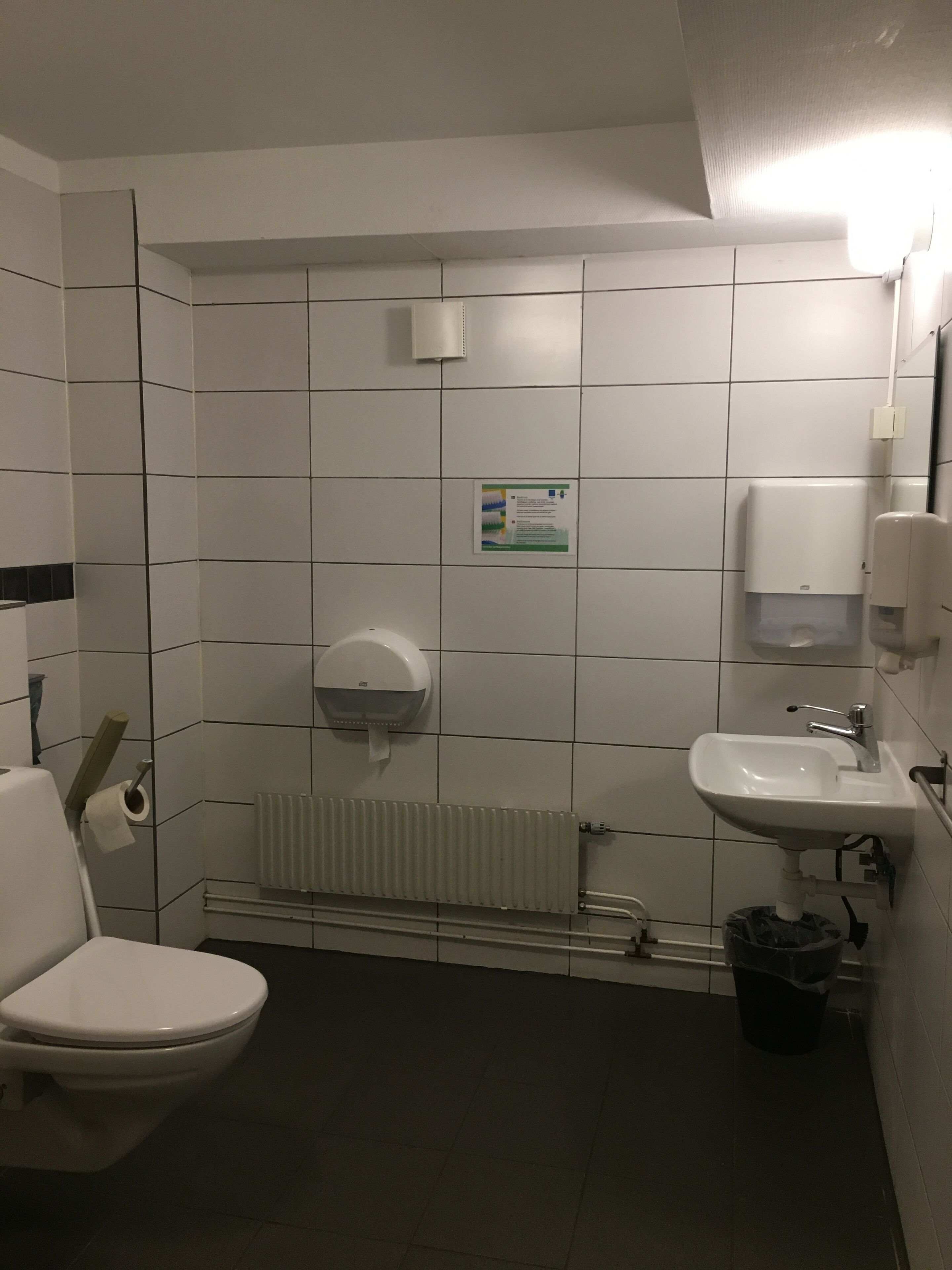 Pieni, siisti kylpyhuone, jossa on valkoiset laatat, wc-istuin, pesuallas, paperipyyheannostelija ja patteri.