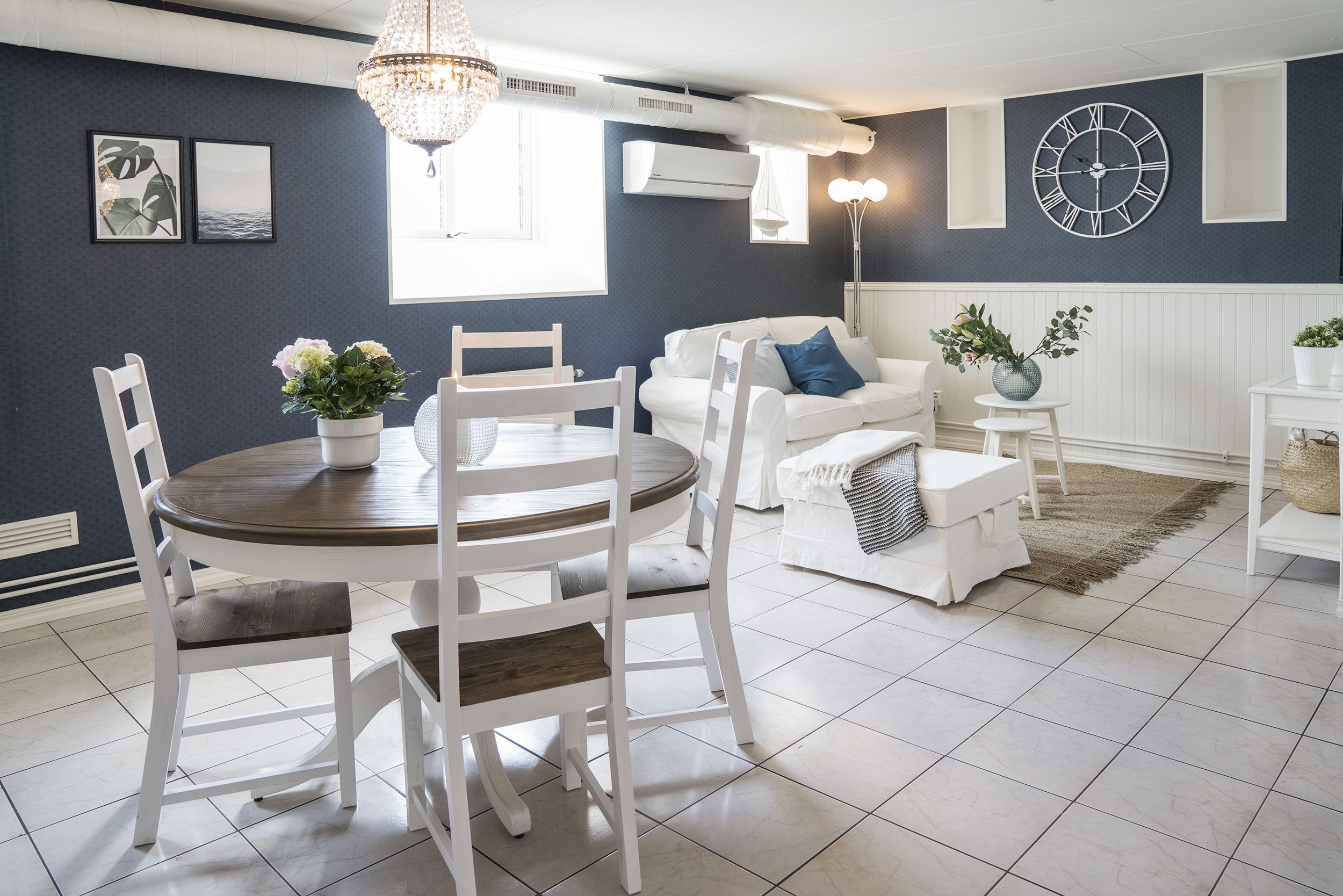A modern living room with a dining area, featuring a chandelier, air conditioning units, and a large wall clock.