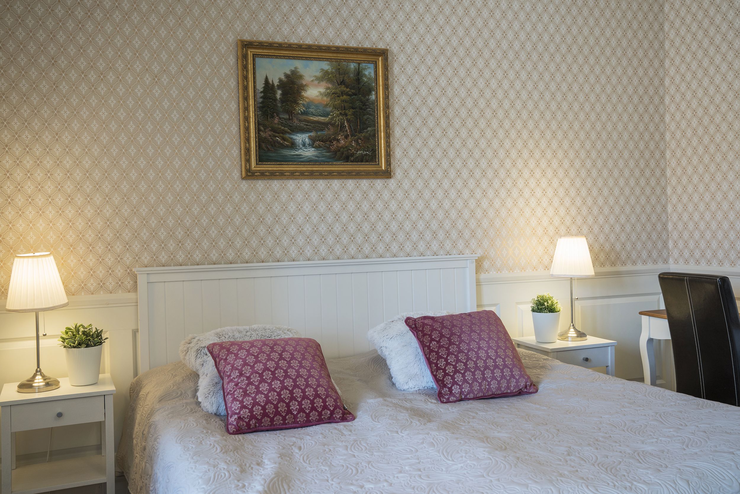 A neatly arranged bedroom with a bed, two nightstands, a chair, and a painting on the wall.
