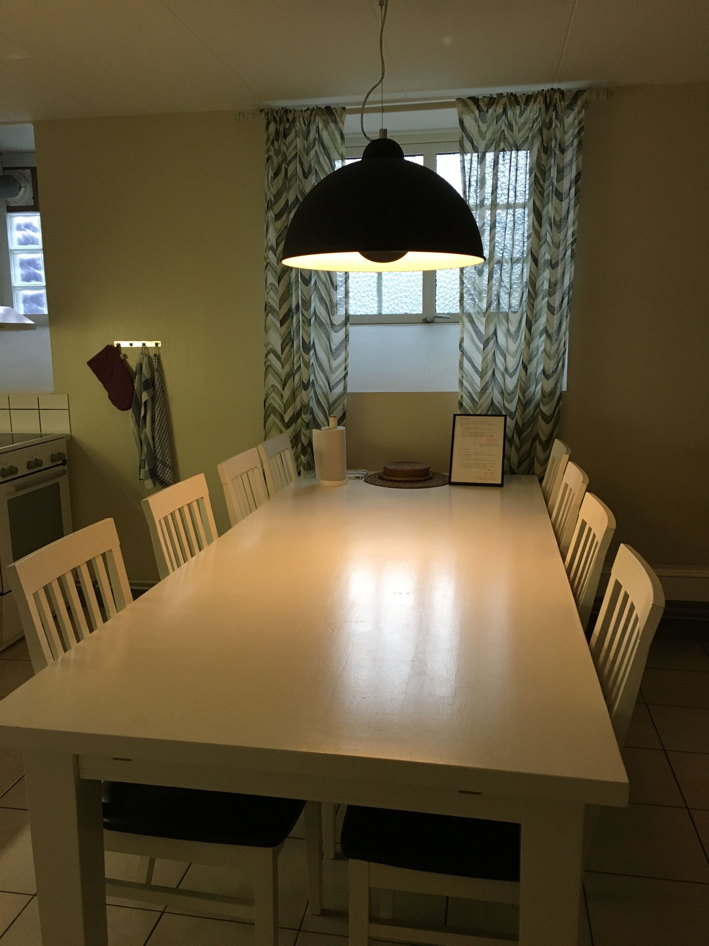 Un comedor con una mesa y sillas blancas, una lámpara colgante, un ventanal con cortinas y una zona de cocina con estufa y utensilios.