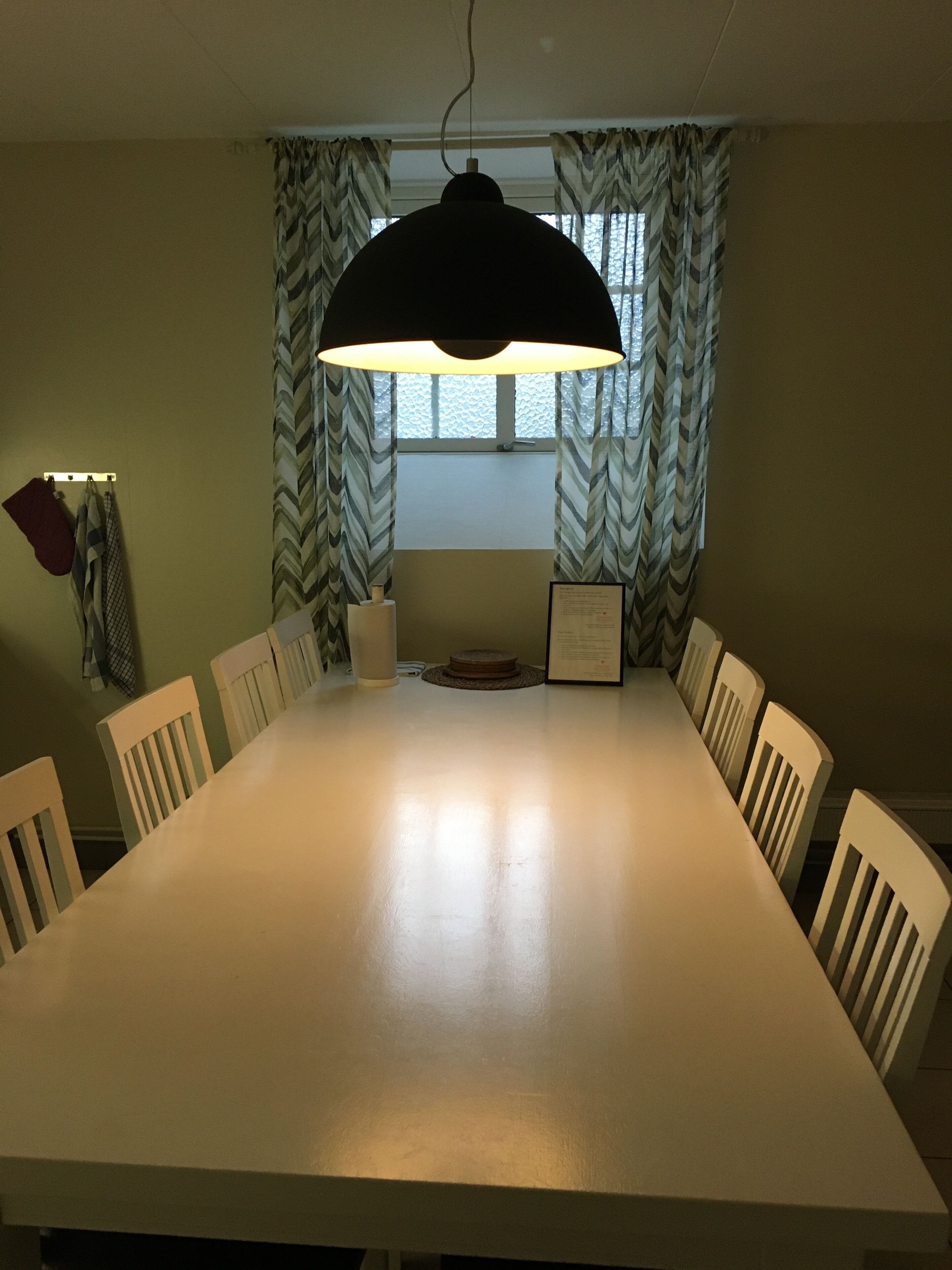 A dining area with a large table, white chairs, a hanging lamp, and patterned curtains.