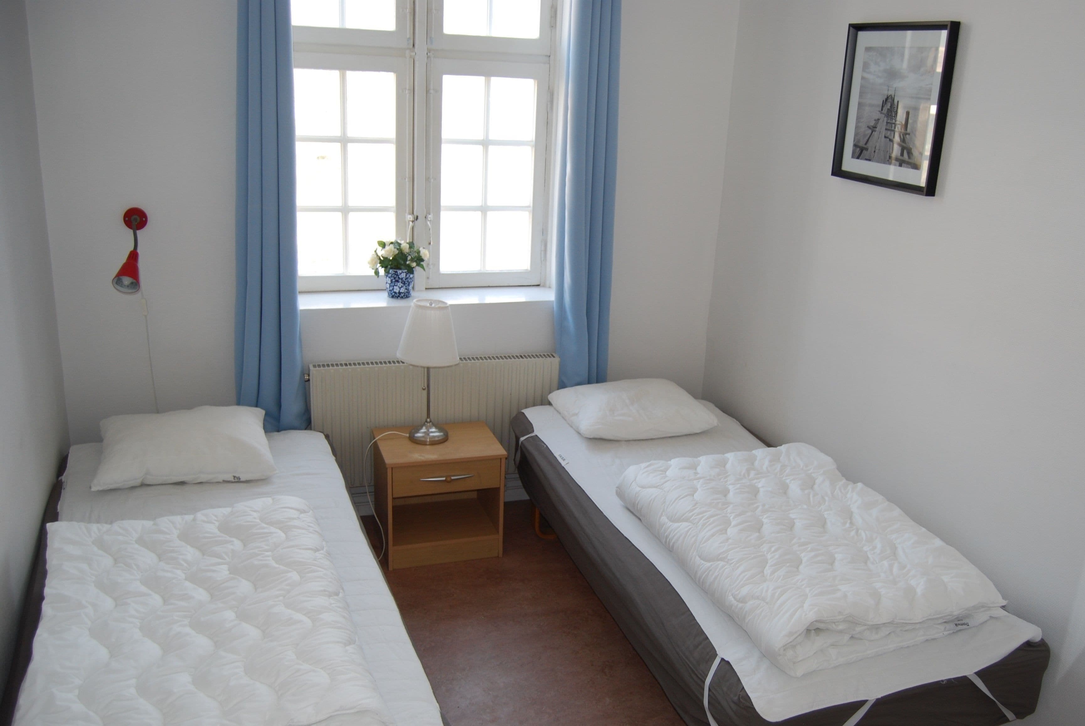 Et værelse med to enkeltsenge, et sengebord med lampe, et vindue med gardiner og et billede i ramme på væggen.