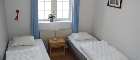 Et værelse med to enkeltsenge, et sengebord med lampe, et vindue med gardiner og et billede i ramme på væggen.