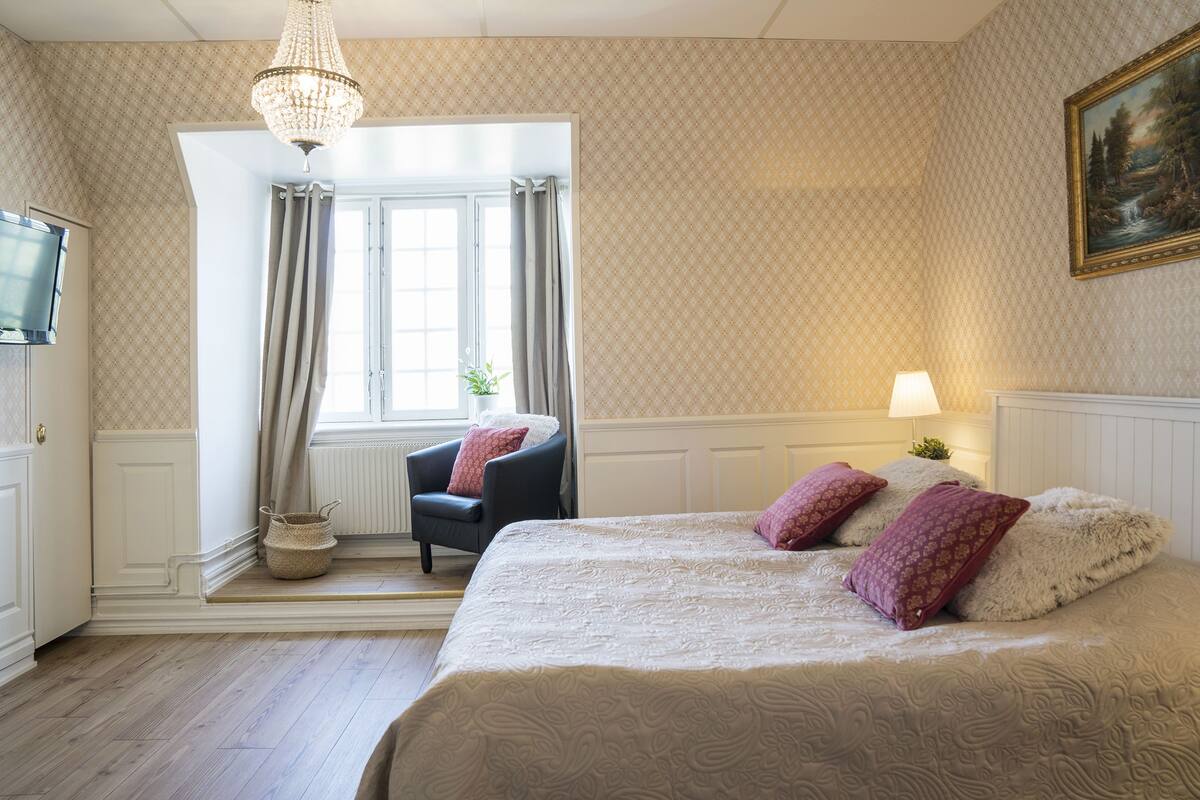 A bedroom with a large bed, a chair, a television, and a window with curtains.