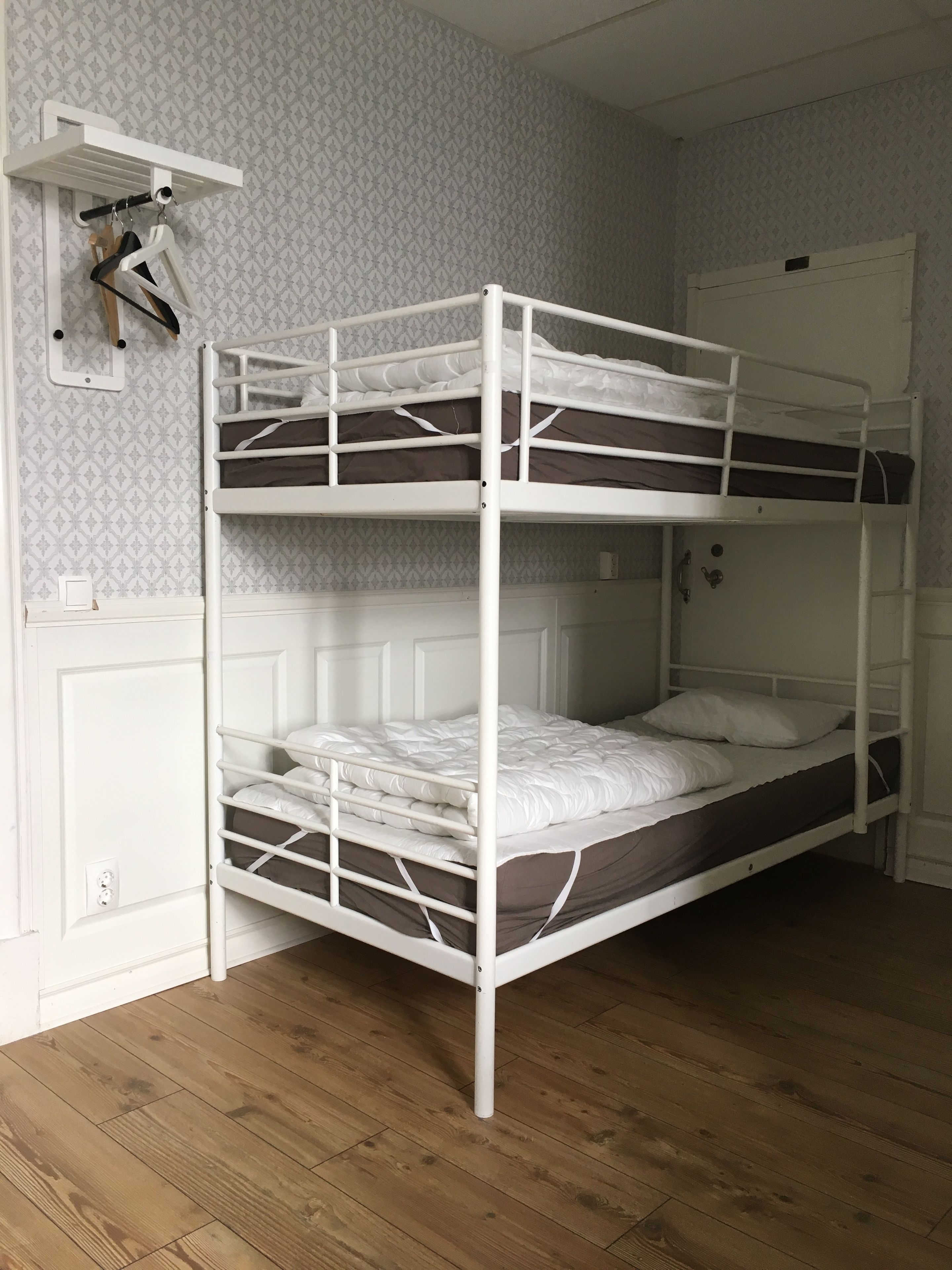 A bunk bed room with a white metal frame, wooden flooring, and patterned wallpaper.