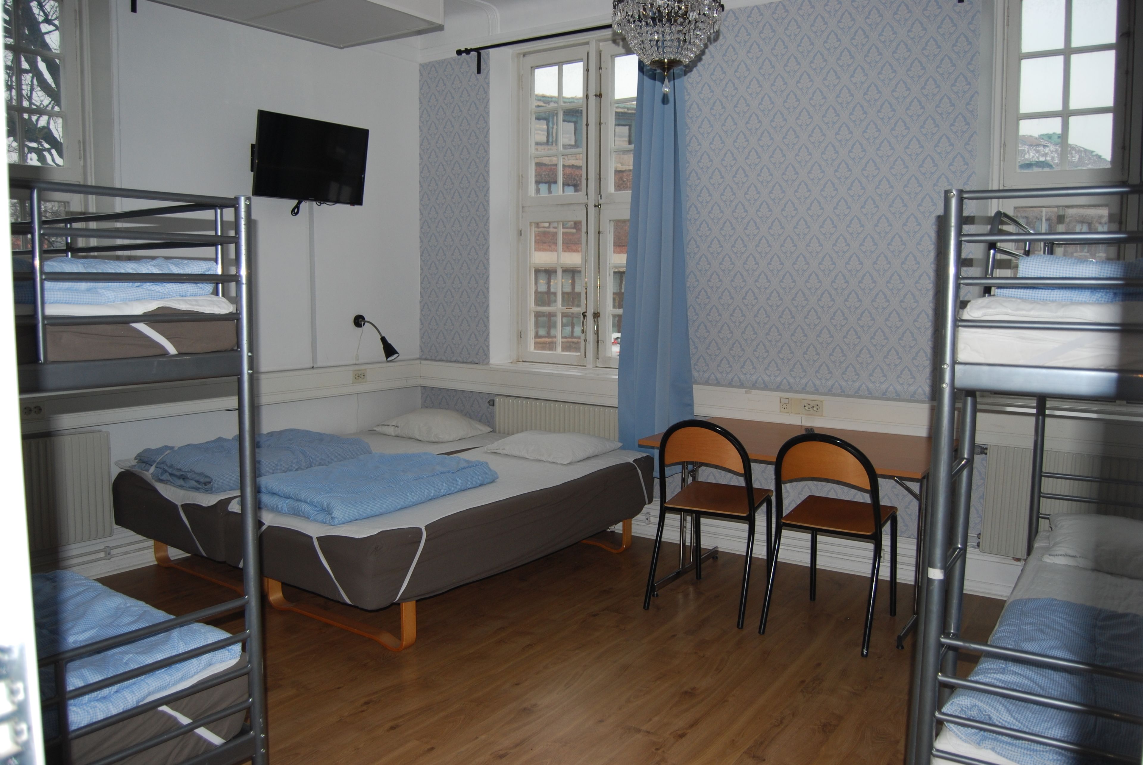 A dormitory room with bunk beds, a desk, two chairs, a television, and a window with a curtain.