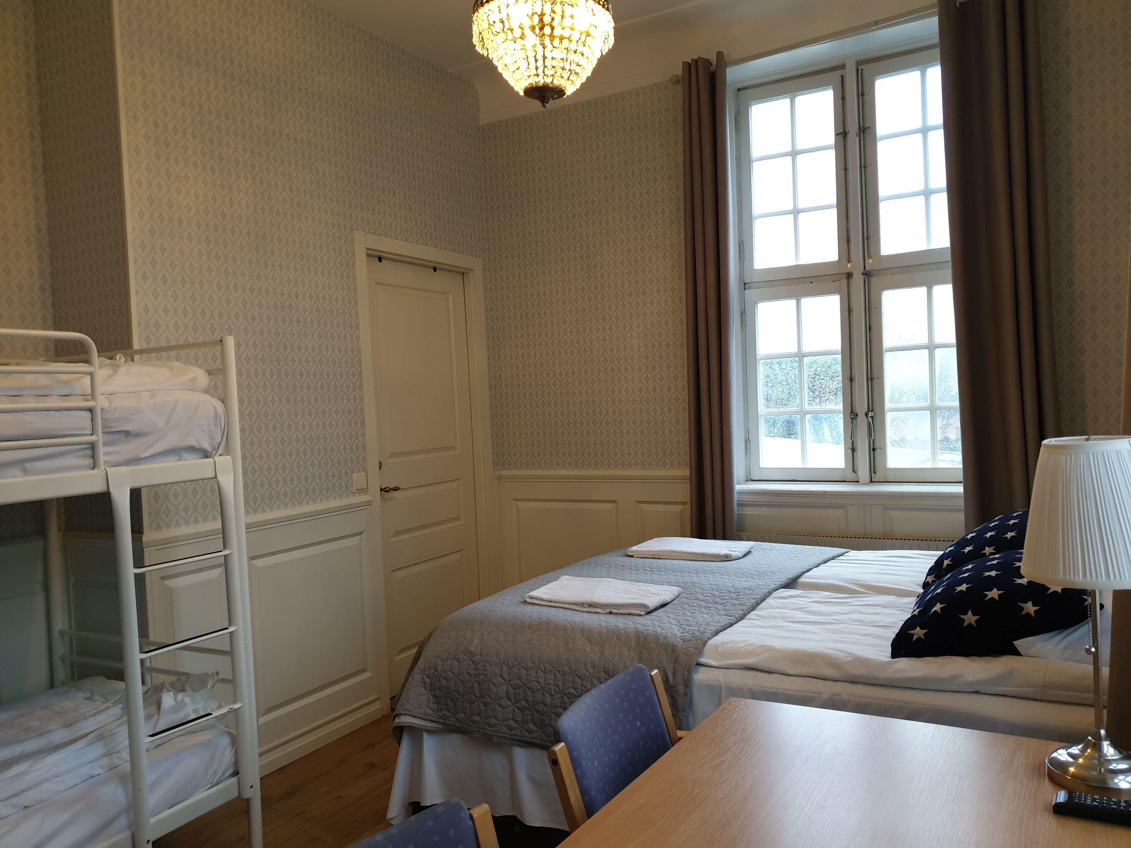 Hotelkamer met een stapelbed, een enkel bed, een bureau met lamp en een raam met gordijnen.