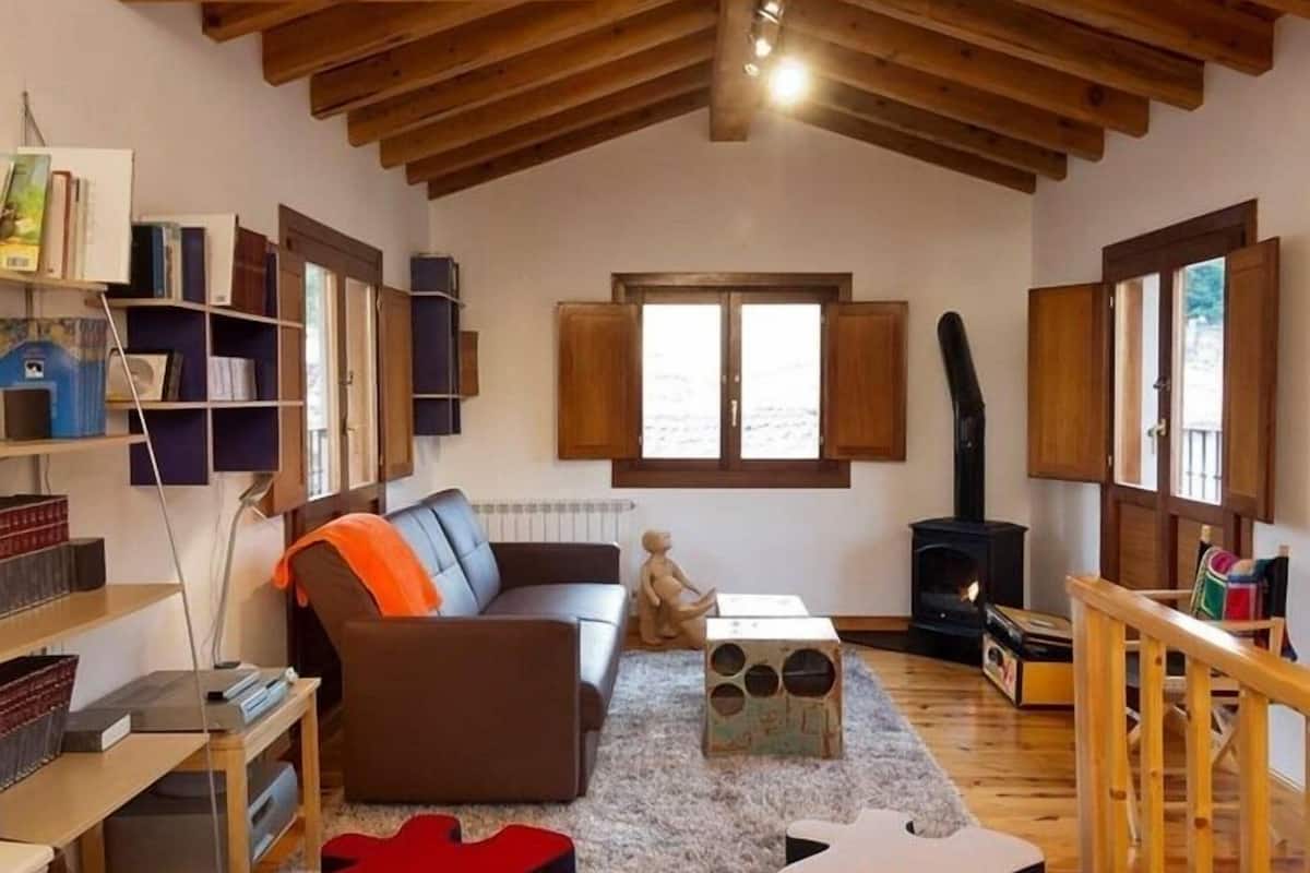 A cozy living room with a high wooden ceiling, a fireplace, a sofa, a bookshelf, and a window with wooden shutters.