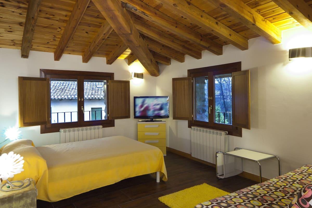 A bedroom with a wooden ceiling, a bed, a TV, and windows with shutters.