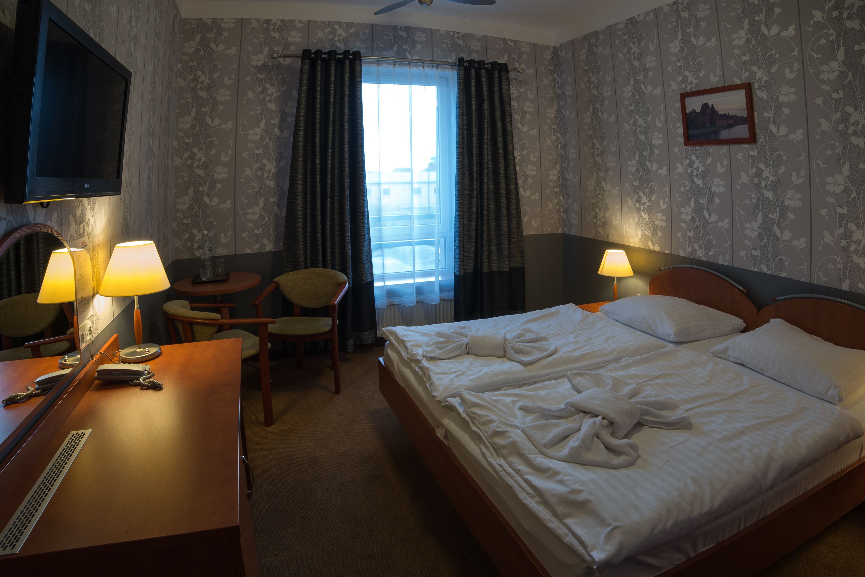 Una habitación de hotel con una cama, dos lámparas de noche, un escritorio con teléfono, una silla y un televisor montado en la pared.