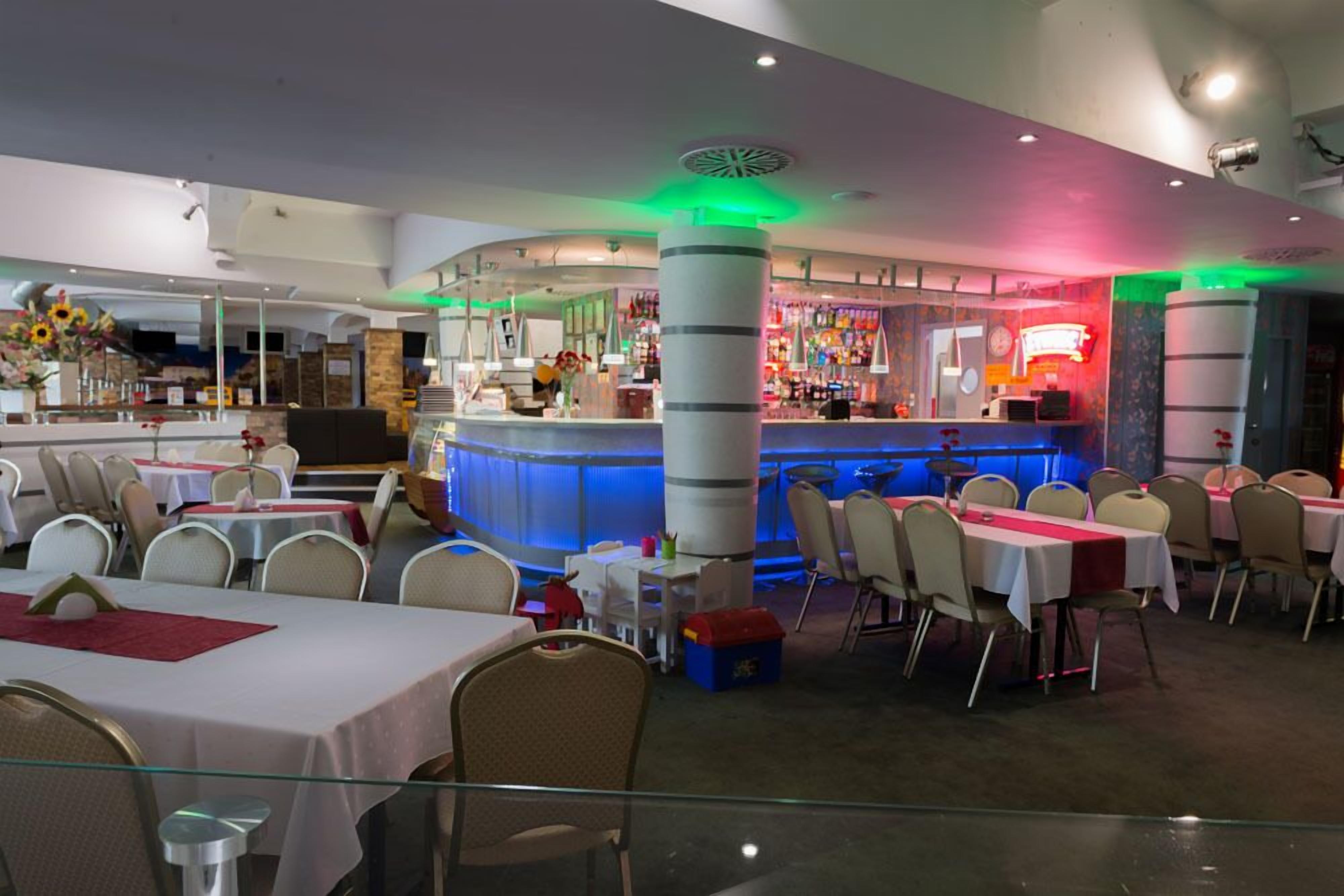A restaurant with multiple tables and chairs, a bar area with colorful lighting, and a decorative column.
