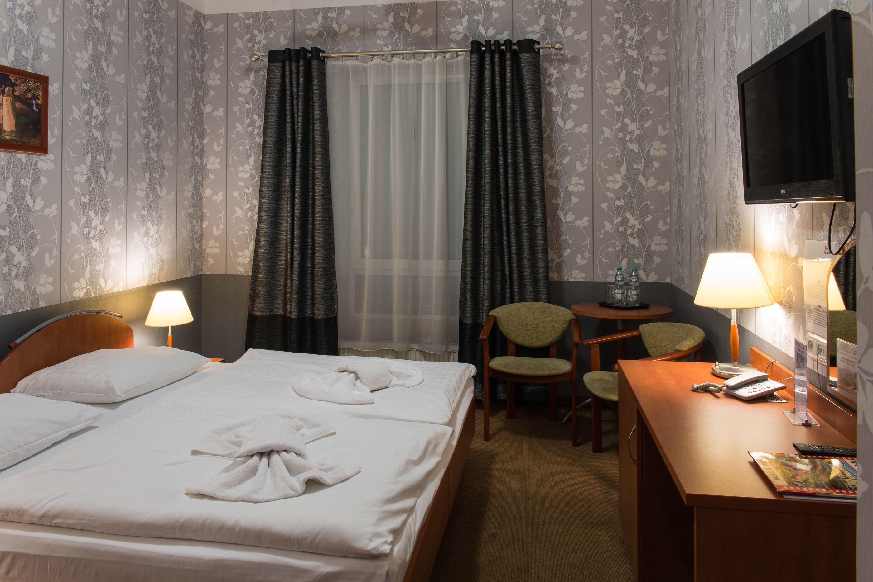 A hotel room with a bed, two chairs, a desk, a TV, and a window with curtains.