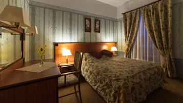 A hotel room with a wooden desk, two bedside lamps, a bed with floral bedding, a chair, and a window with curtains.